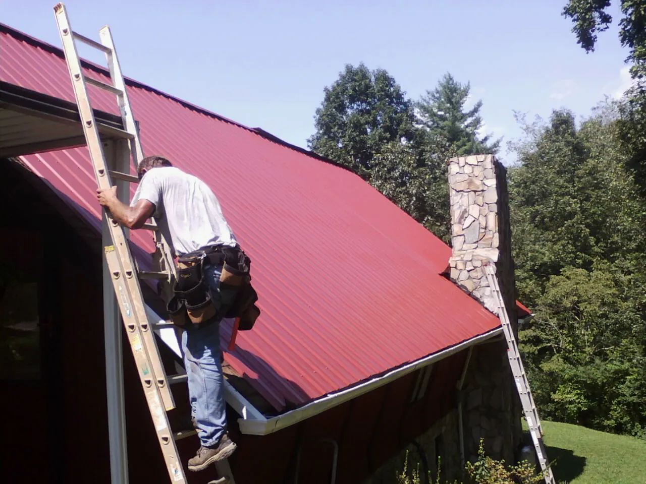 Roofer on a ladder beside a red metal roof and stone chimney — Soffit & Fascia Repair by Roofing Brothers in Basin