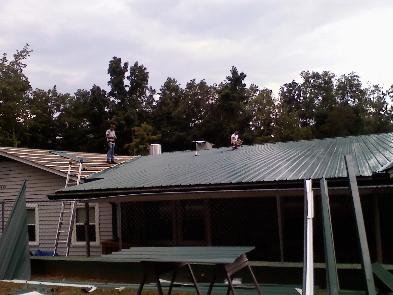 Two roofers installing green metal panels above a covered porch in Basin